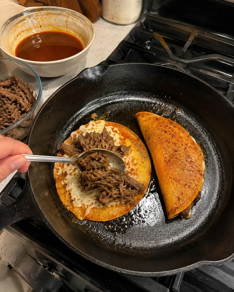 The Crockpot Birria Taco being filled with shredded beef and cheese in a skillet
