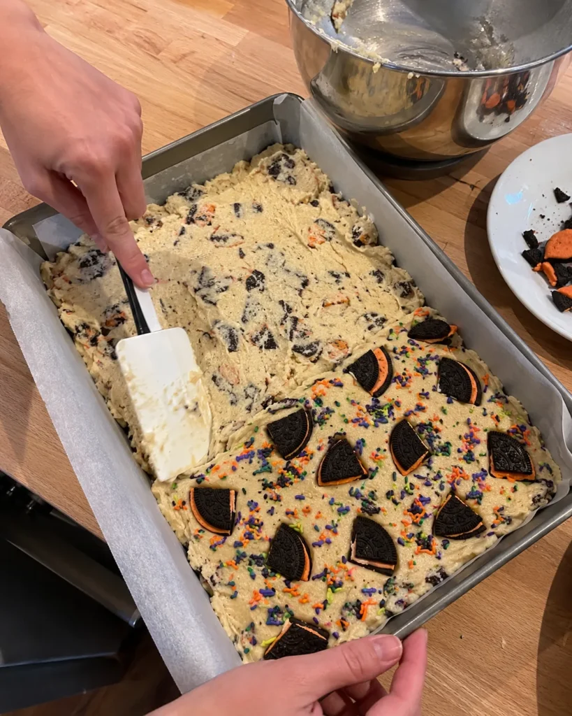 Thick Halloween Oreo Cake Bars dough being pressed into a parchment-lined baking pan