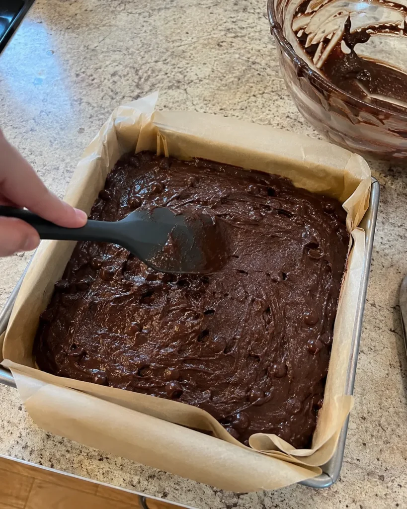 Chocolate zucchini brownie batter being spread into a parchment-lined baking pan